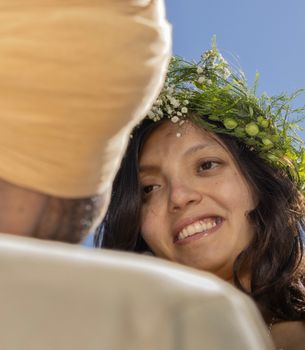 A joyful bride adorned with a floral crown smiles widely during an outdoor wedding ceremony under a clear blue sky.