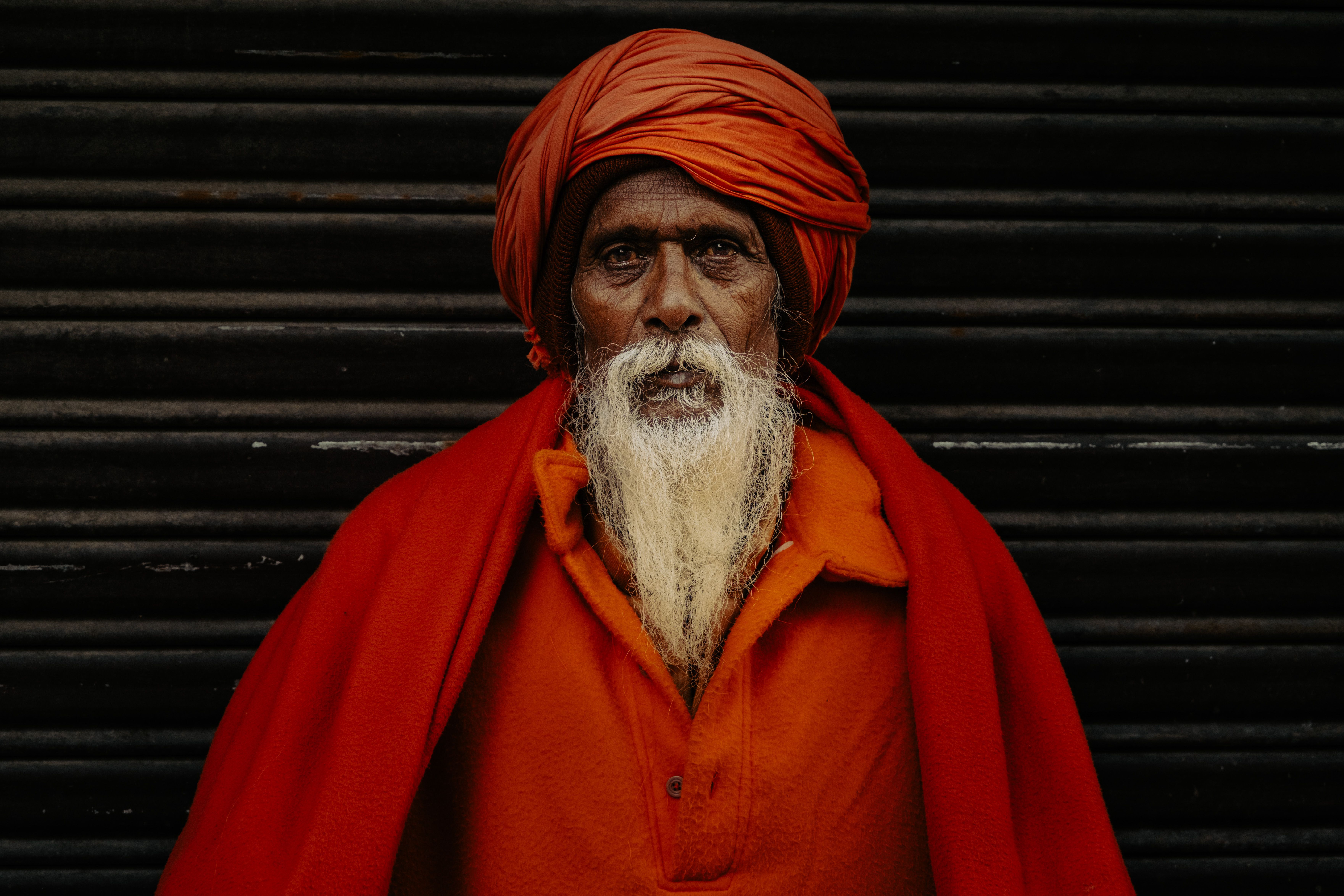 Elderly man with white beard in bright orange robe and turban stands against a dark background.