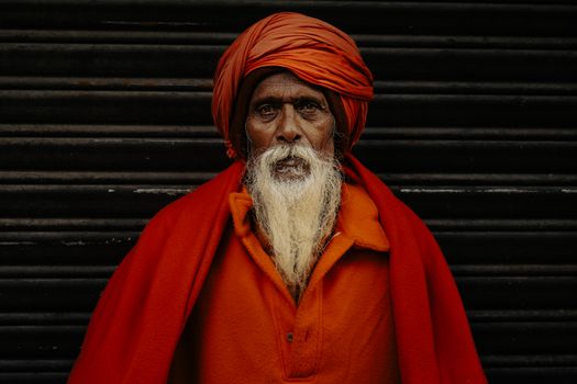 Elderly man with white beard in bright orange robe and turban stands against a dark background.