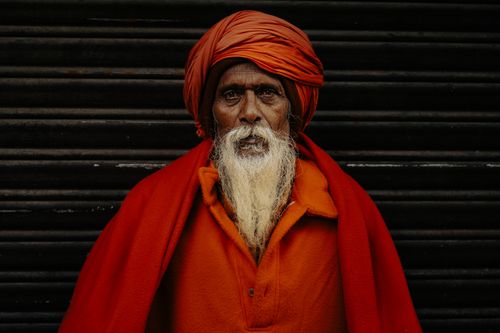 Free Elderly man with white beard in bright orange robe and turban stands against a dark background. Stock Photo