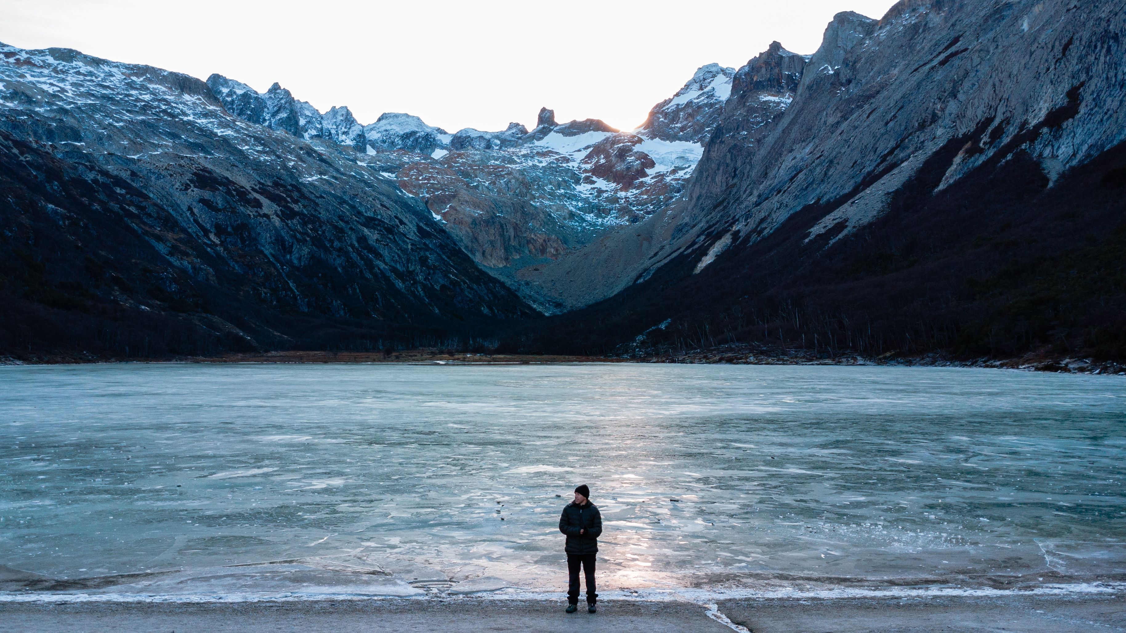 A lone figure on a dark shore facing a glacier and steep snow-streaked mountains.