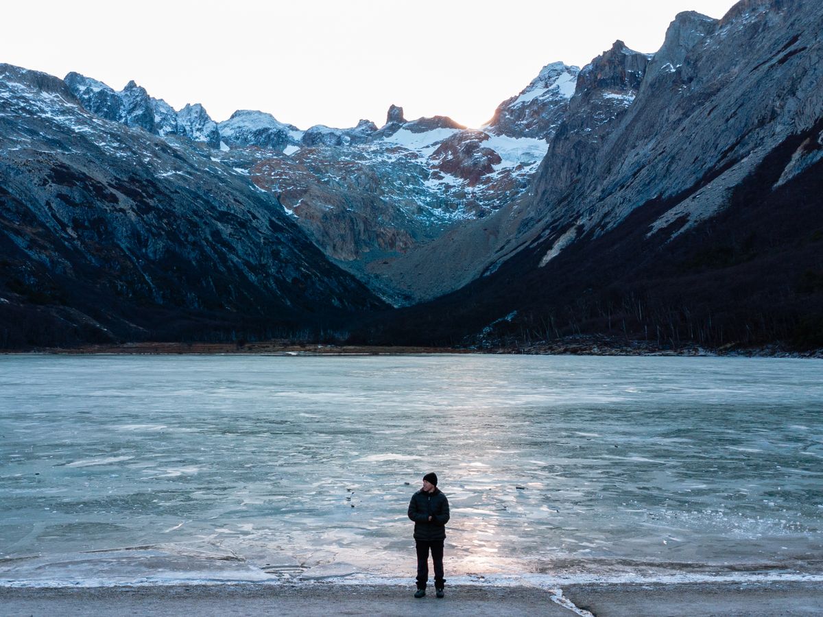 A lone figure on a dark shore facing a glacier and steep snow-streaked mountains.