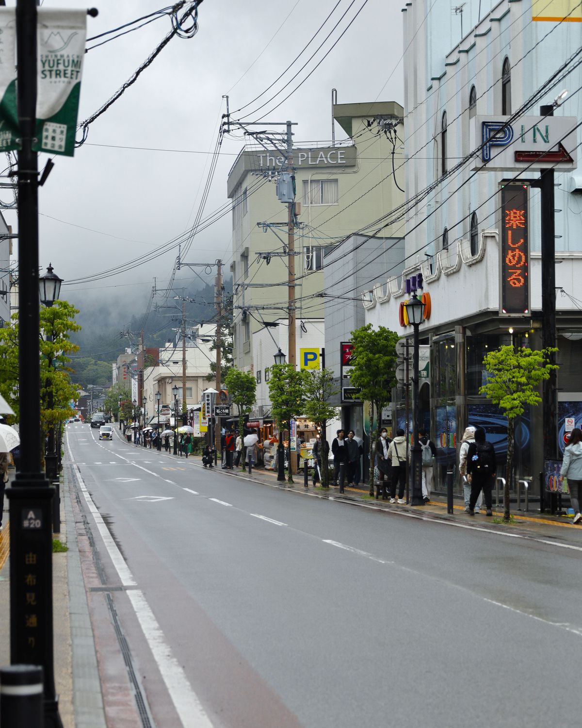 Shop-lined street in Yufu, Oita, with pedestrians walking under umbrellas on a misty, rainy day