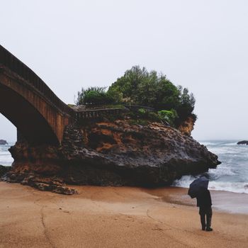 A lone figure with an umbrella stands on a rainy beach in Biarritz, France, by the rocky shore and a bridge.