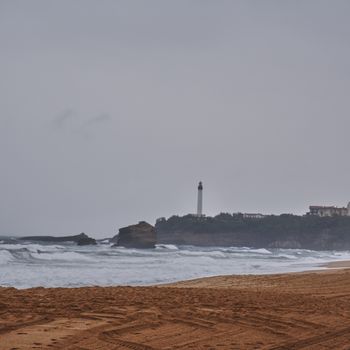 A serene view of the Biarritz lighthouse and beach under overcast, stormy skies with crashing waves.
