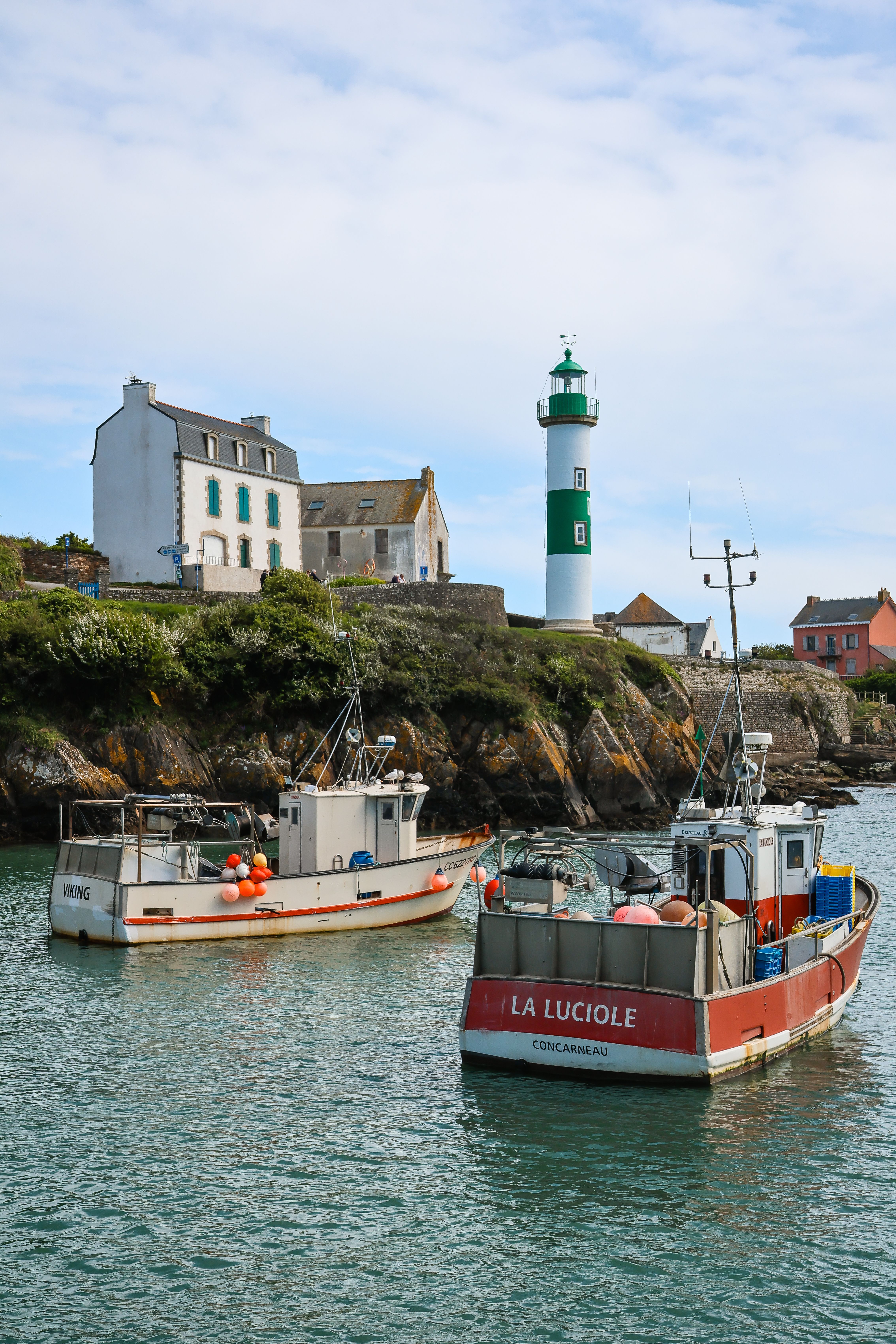 Two fishing boats anchored near a French coastal lighthouse and houses, capturing a peaceful scenery.