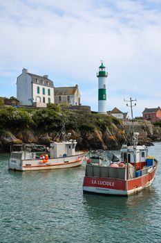 Two fishing boats anchored near a French coastal lighthouse and houses, capturing a peaceful scenery.