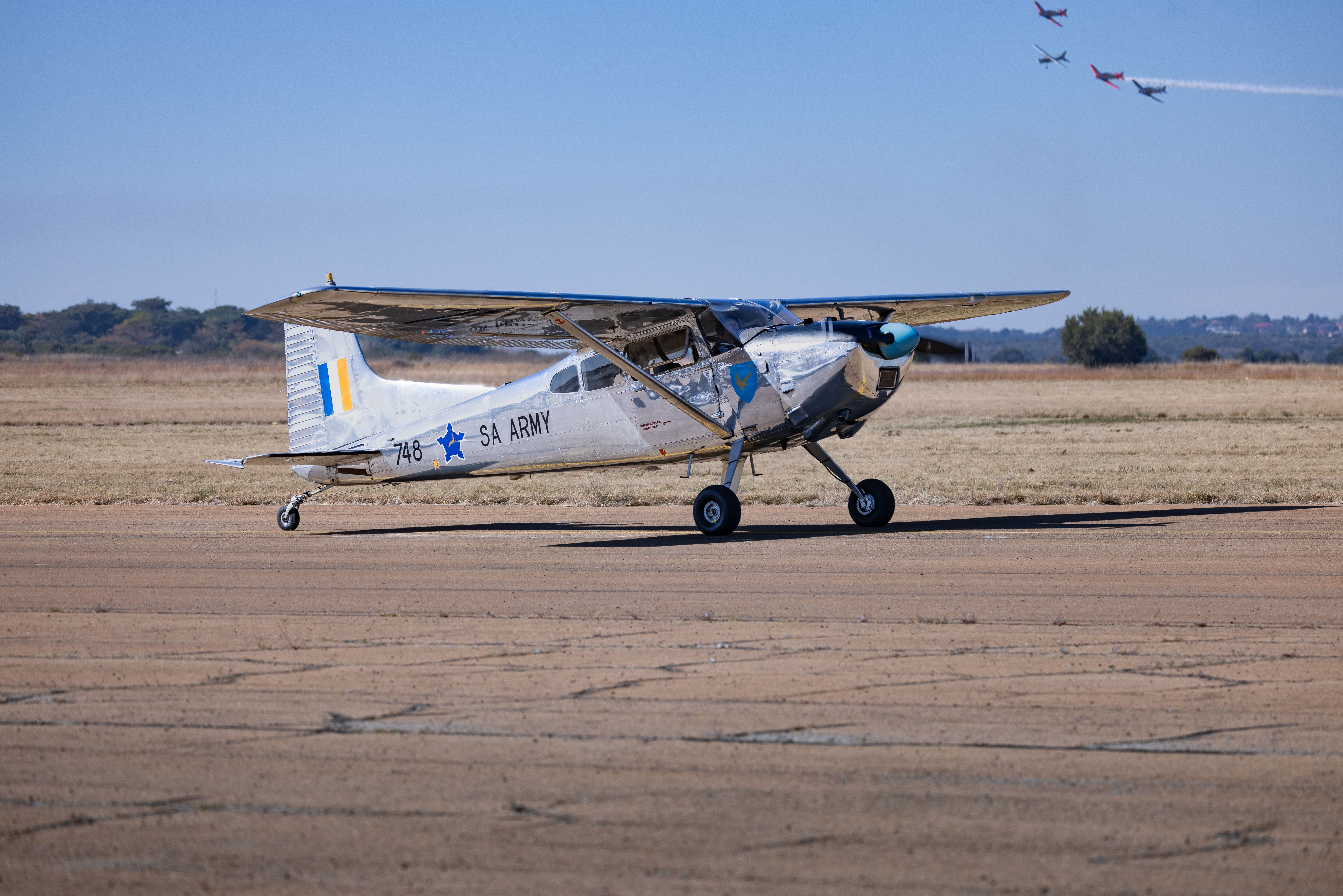 A classic SA Army airplane captured on the runway with a formation of jets in the sky.