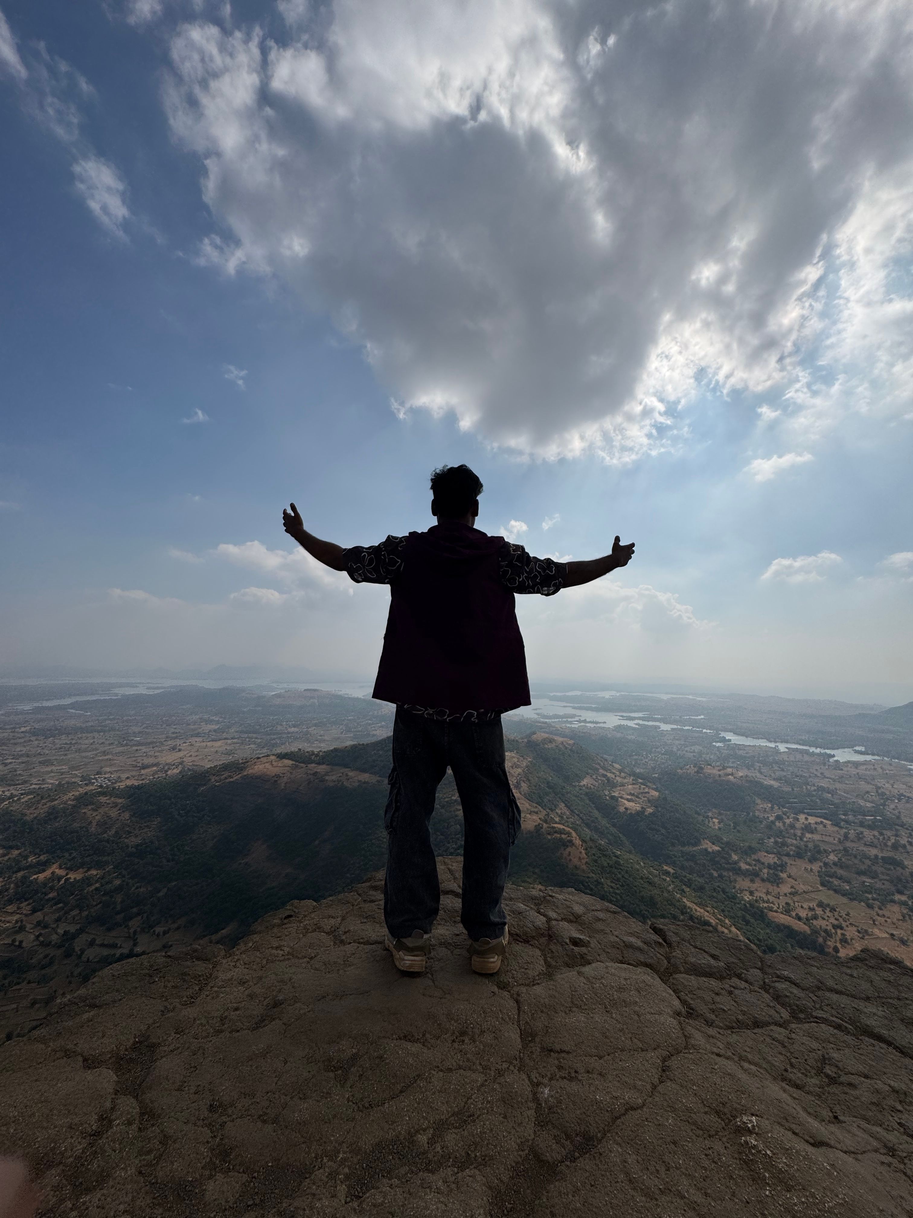 A person stands on a mountain peak in Mumbai, arms outstretched, embracing a vast scenic view under a cloud-filled sky.