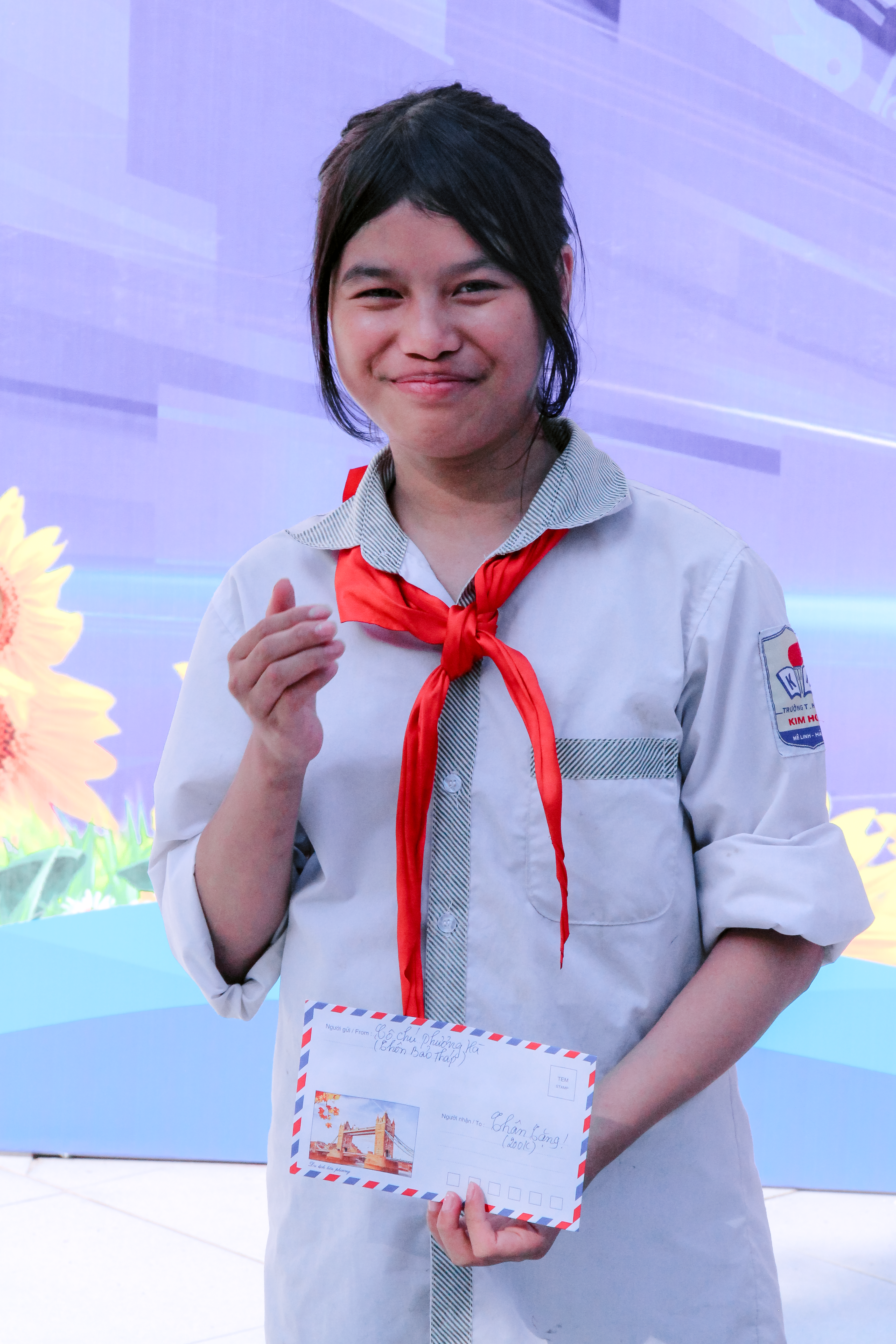 A smiling student in a uniform holds an envelope confidently outdoors.