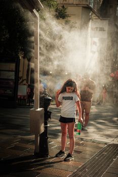 A girl stands under a misting fan on a sunny day in the city, cooling off.