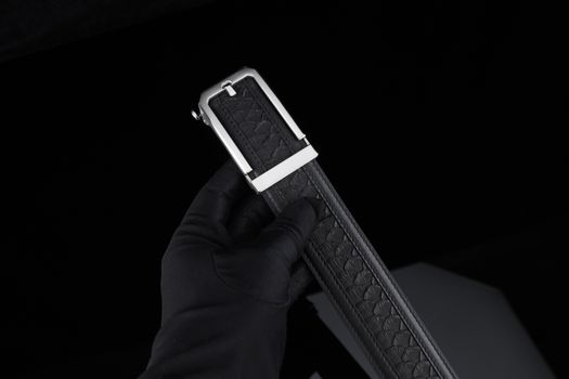 Elegant black leather belt with embossed design and silver buckle held by gloved hand.