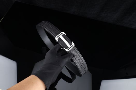 Stylish black leather belt with chrome buckle held by a gloved hand, showcasing luxurious texture.