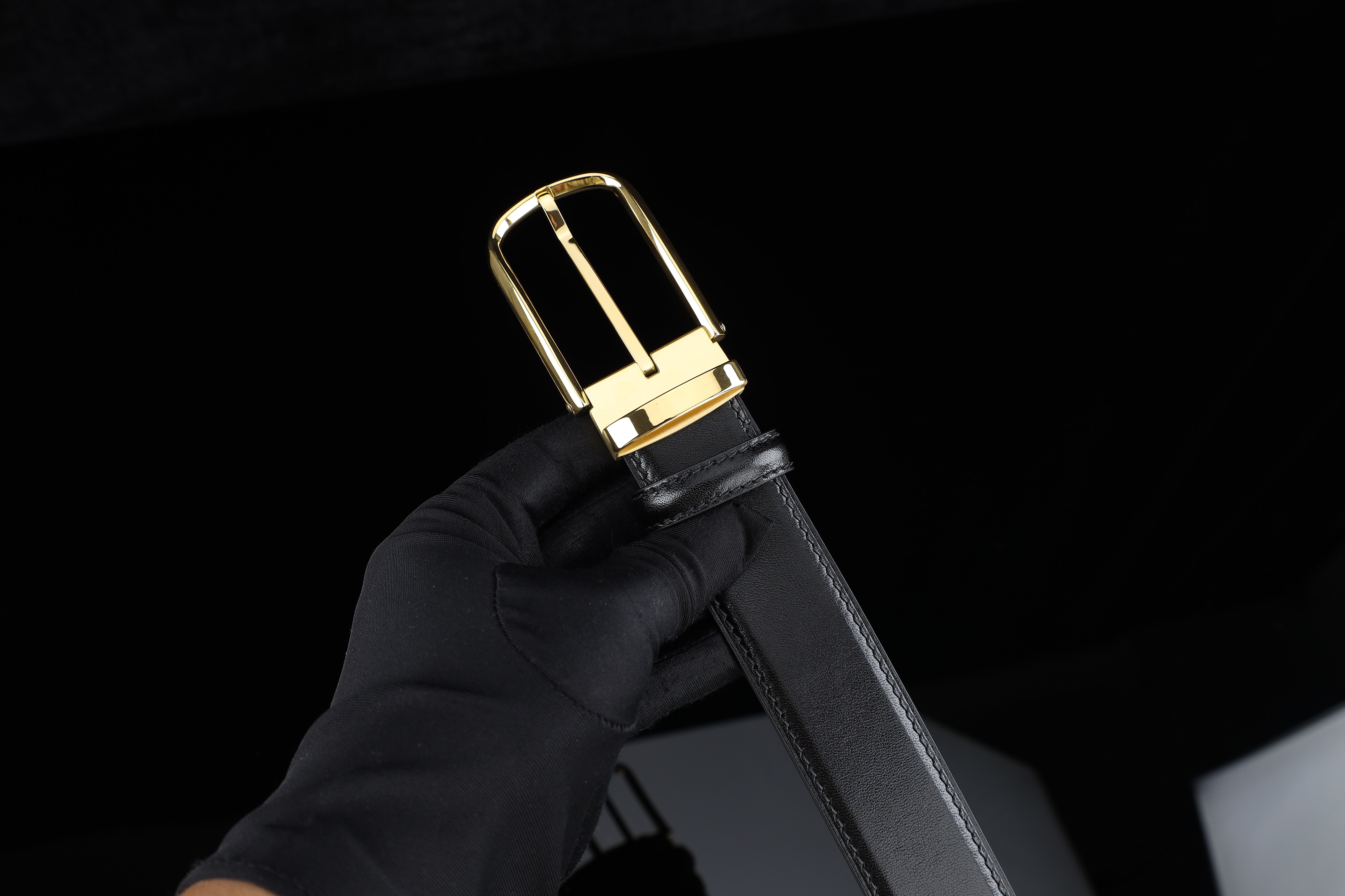 Close-up of a luxury leather belt with a sleek gold buckle, held by a gloved hand on a dark backdrop.