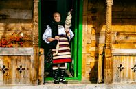 Elderly woman in traditional Romanian clothing standing in a rustic wooden doorway.