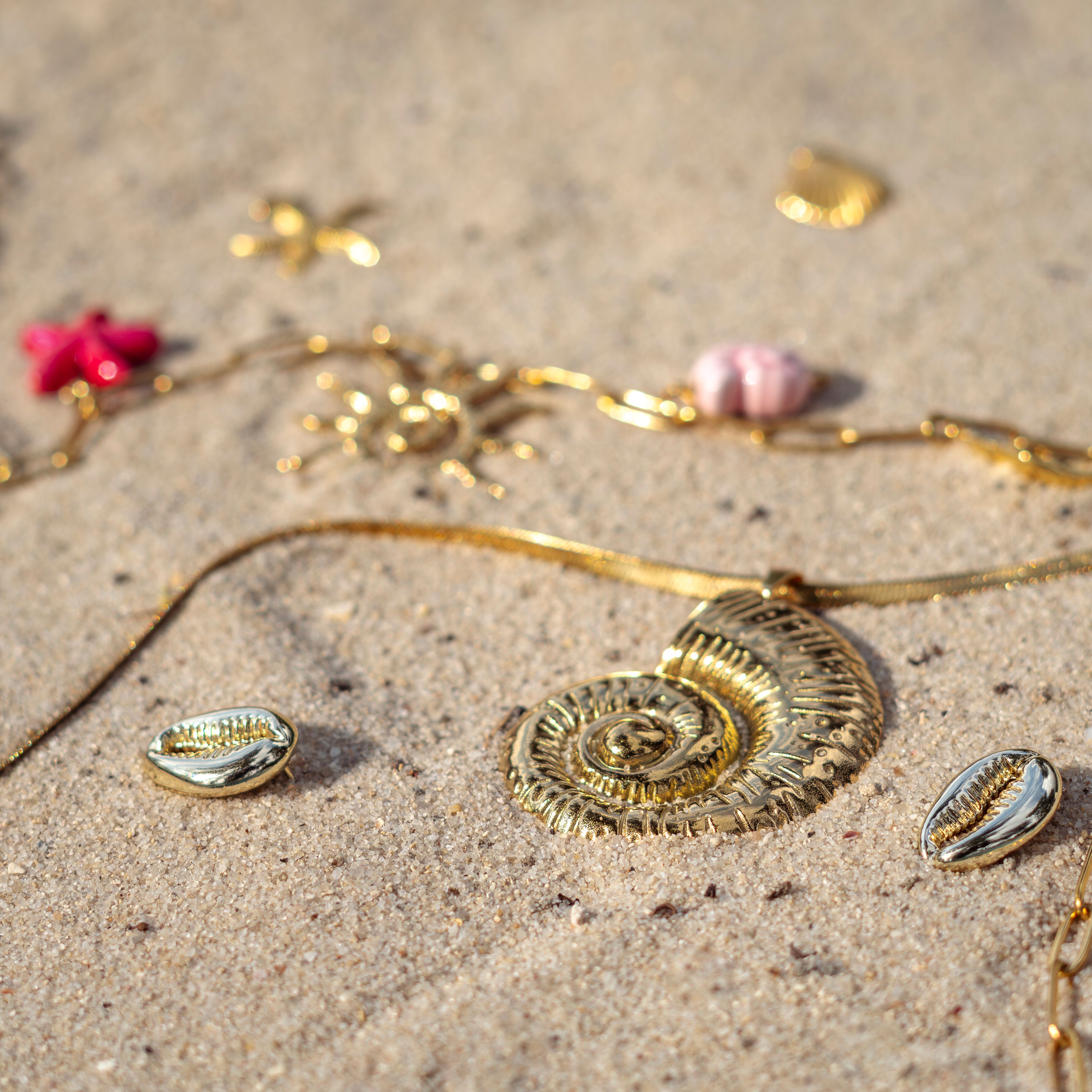 Gold jewelry with seashell designs scattered on the beach sand captured in bright sunlight.