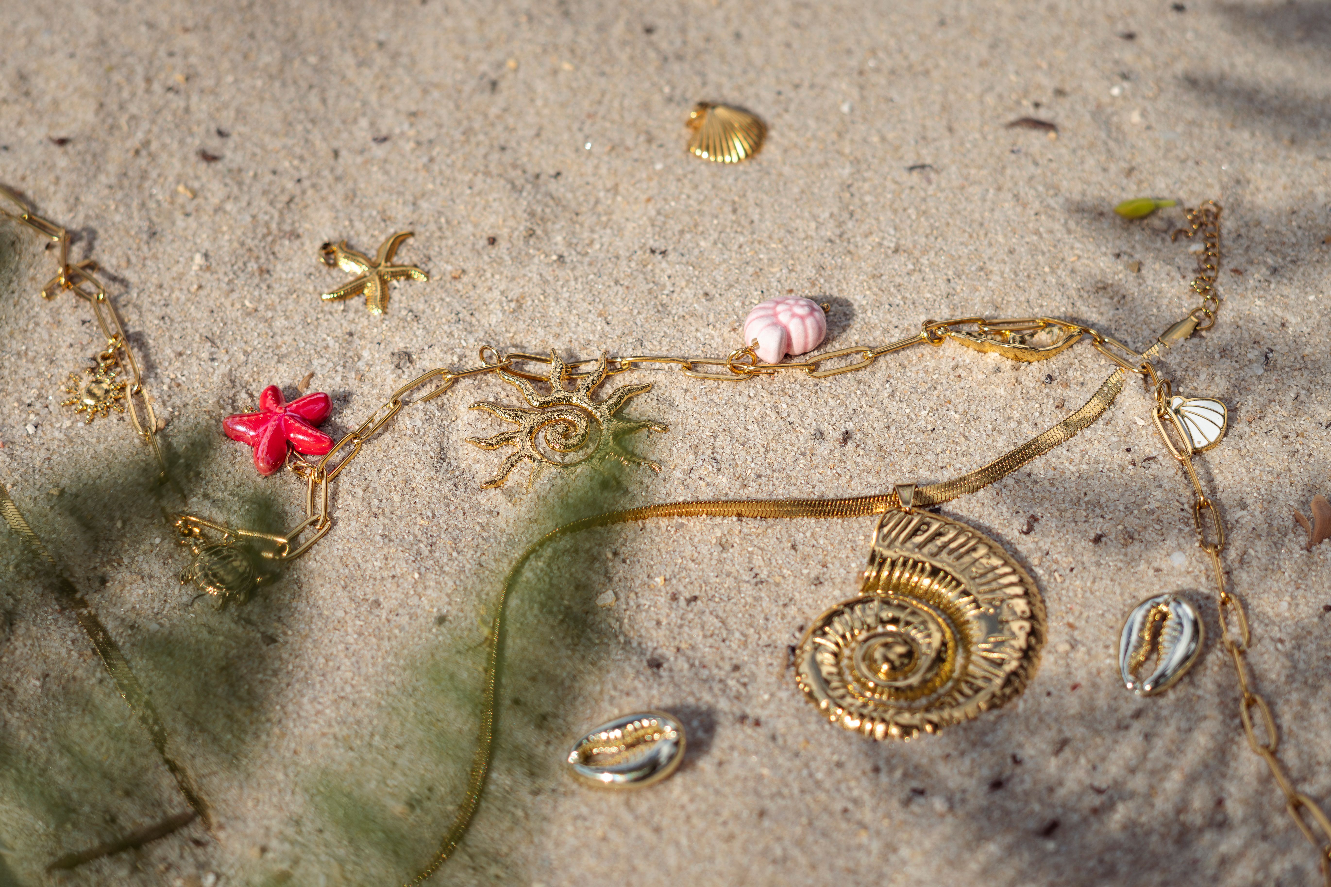 Artistic arrangement of gold jewelry on beach sand, featuring shells and starfish motifs.