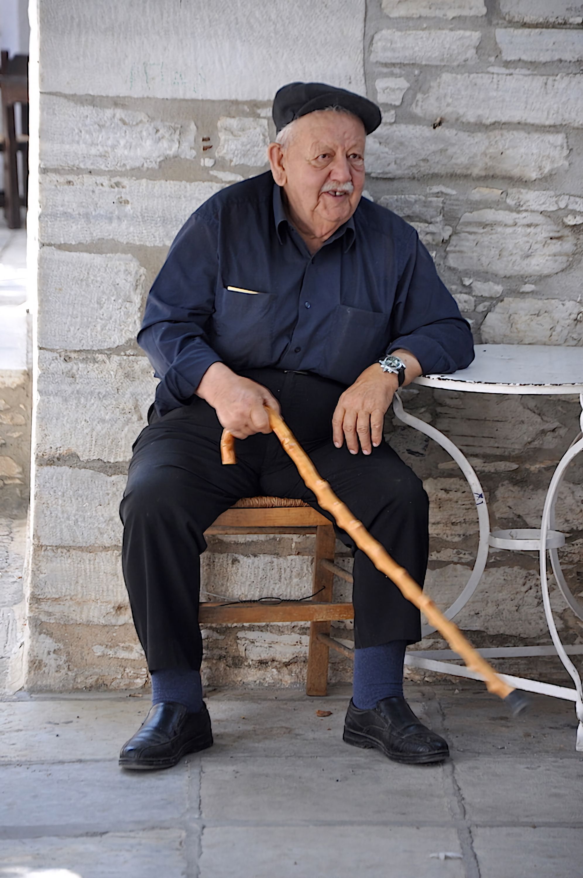 A senior man with a cane enjoys a quiet moment seated outdoors beside a stone wall in a Greek village.