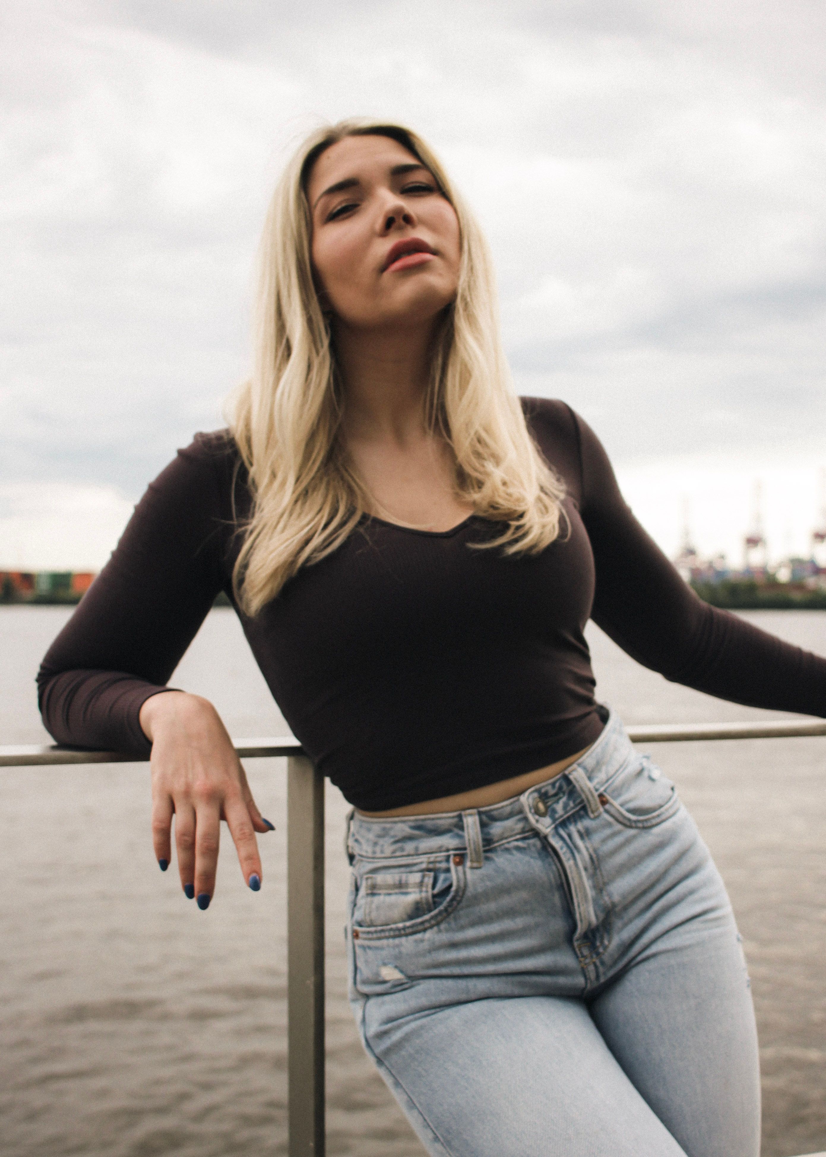 Stylish young woman posing by the Hamburg Harbor on a cloudy day, showcasing casual fashion.