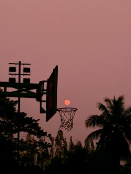 Silhouette of basketball hoop against pink sunset sky with palm tree.