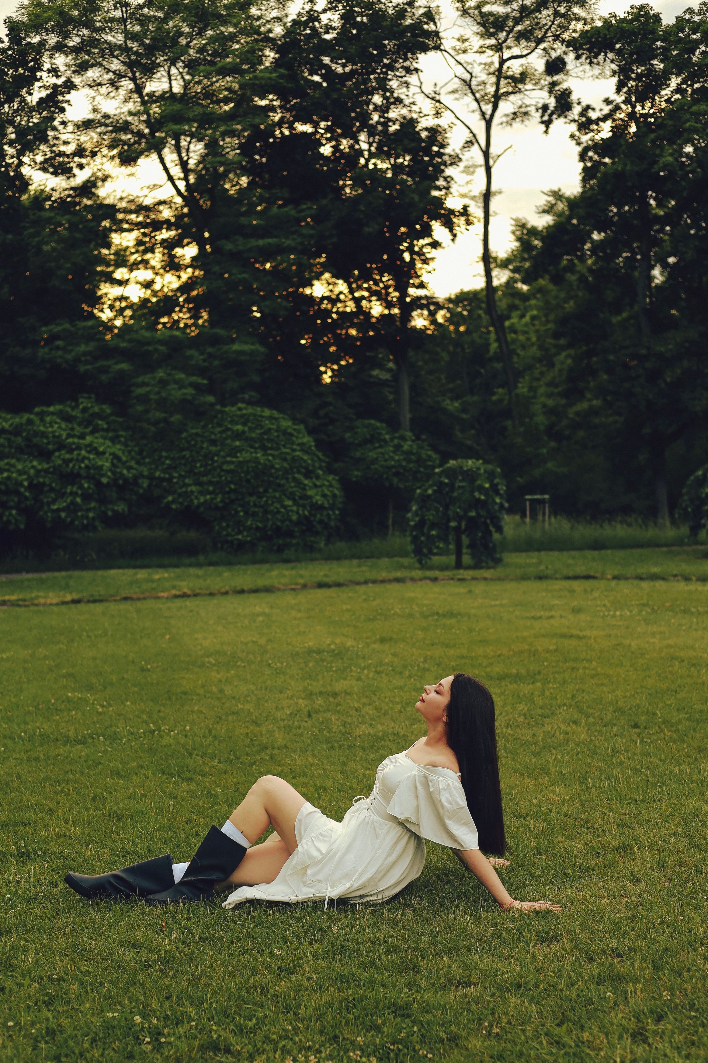 A woman in a white dress relaxes on the grass in a summer park at sunset.