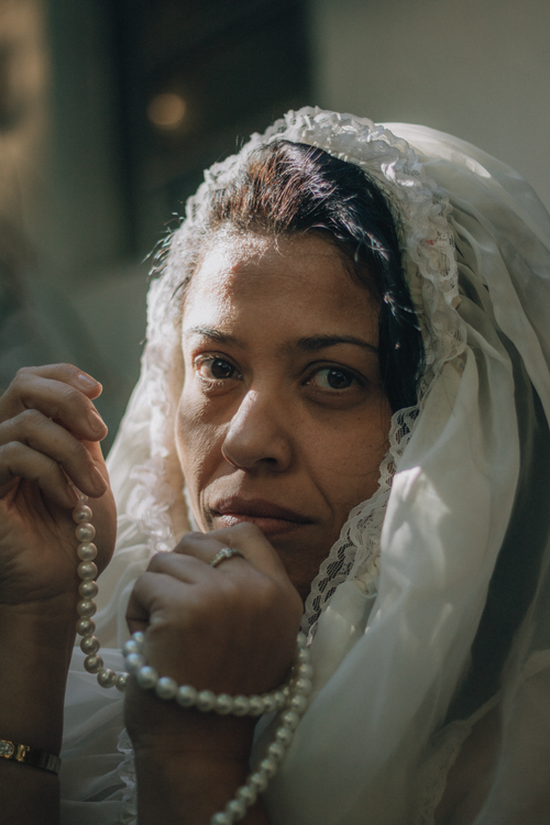 Free A contemplative woman draped in a white veil gently clasping pearl beads in warm light. Stock Photo