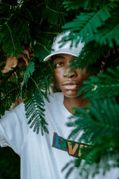 Pensive young African American male millennial in t shirt and cap standing among green plants and looking at camera