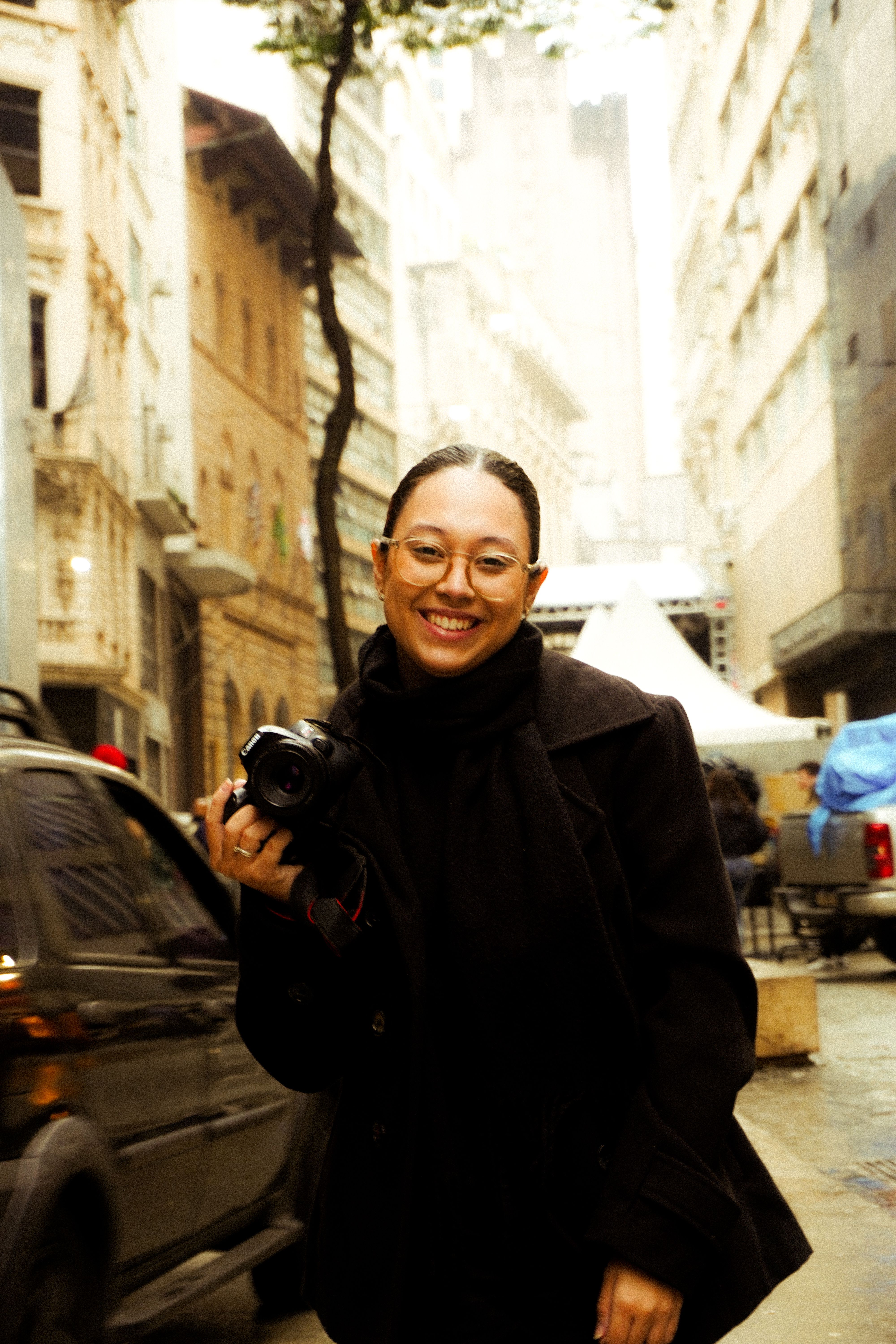 A female photographer smiles while holding a camera in a bustling city street scene.