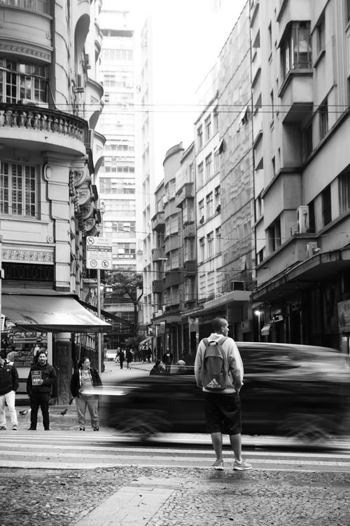 Free Black and white urban street scene capturing motion blur of a passing car and a man standing at a crosswalk. Stock Photo