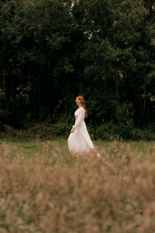 Free A woman in a flowing white dress walks gracefully by a lush green forest. Stock Photo