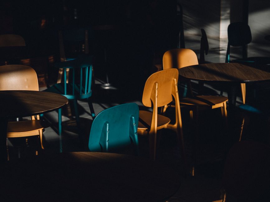 A moody, shadow-filled cafe scene with colorful chairs and tables in dramatic lighting.