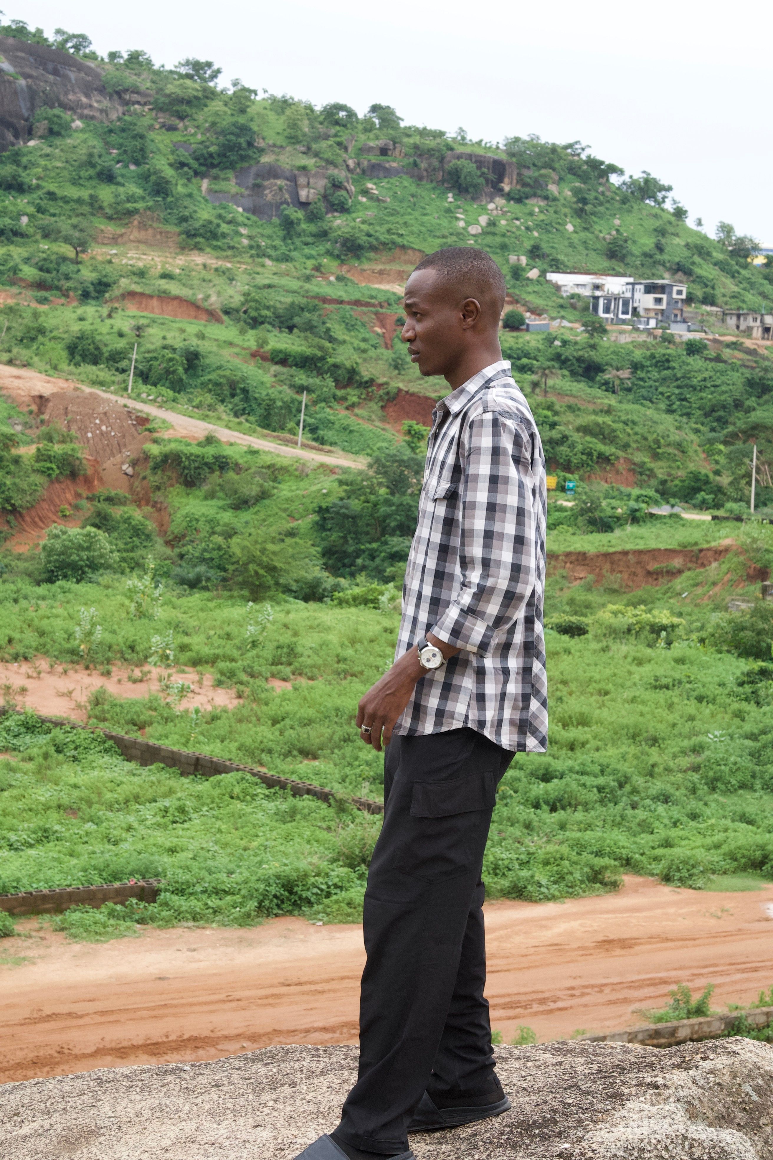A young man in casual wear stands contemplating on a rocky terrain overlooking green landscapes in Abuja, Nigeria.
