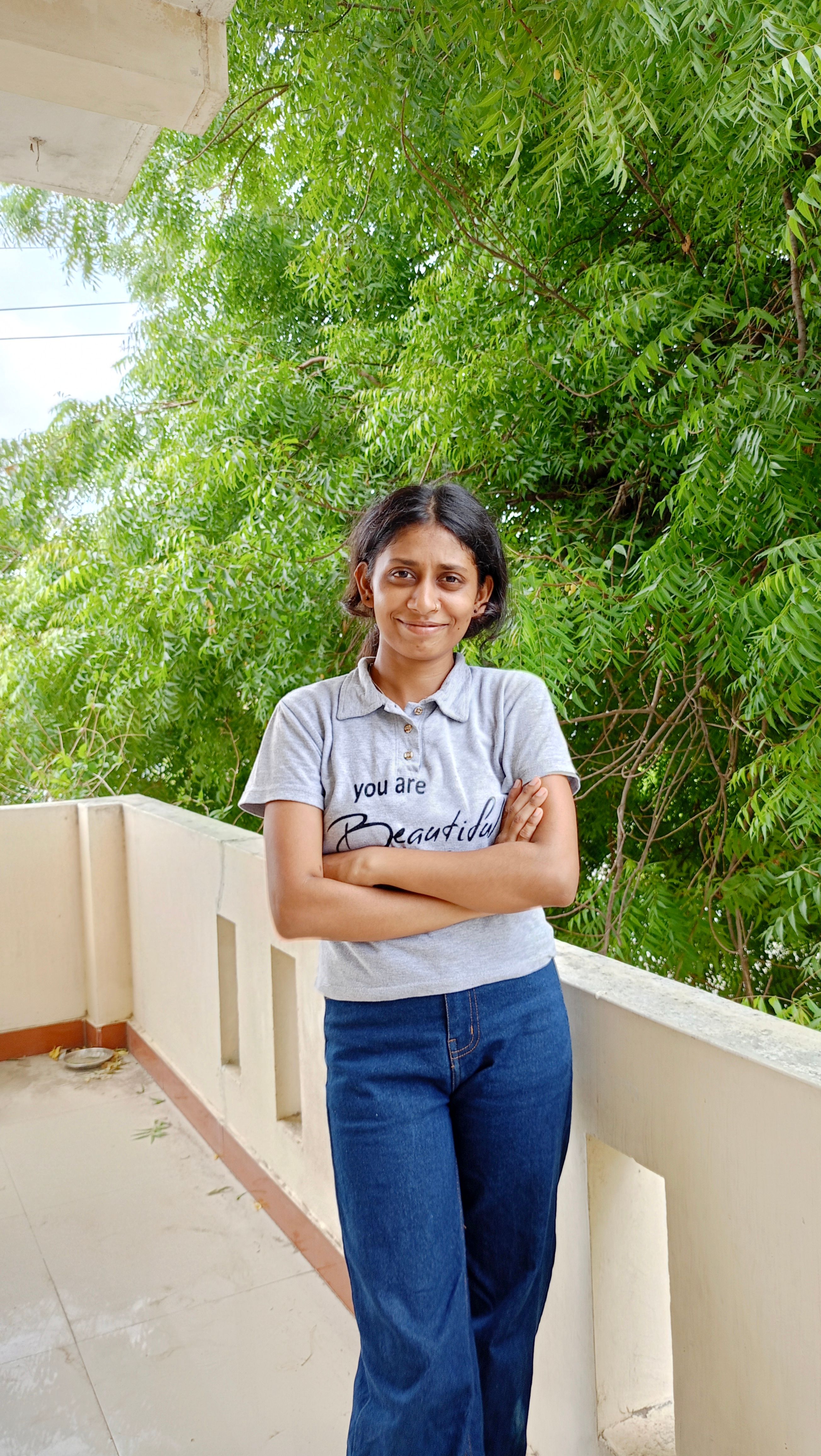 Woman standing on a balcony with lush green trees in the background, smiling confidently.