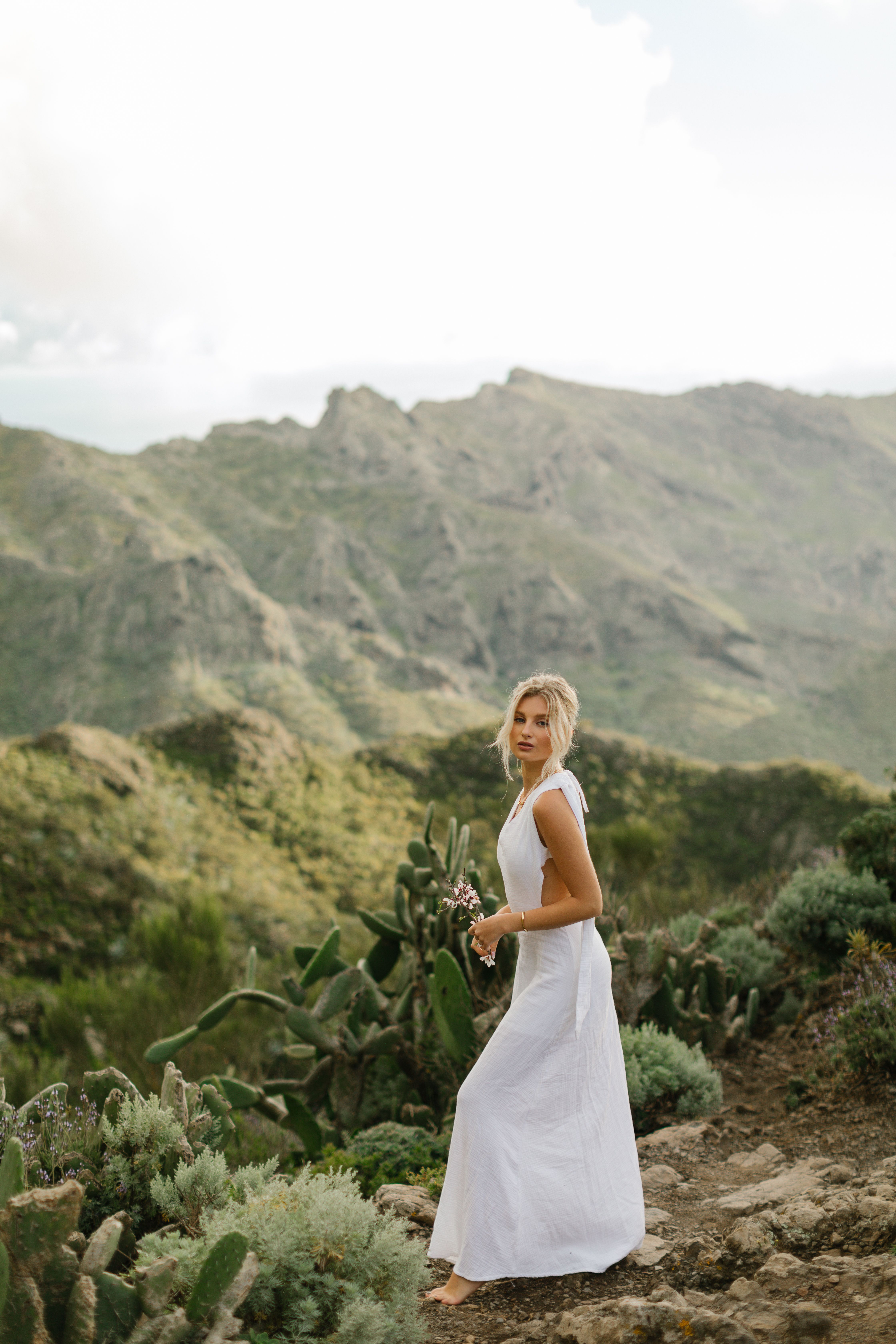 A woman in a white dress stands on a scenic Greek mountain trail surrounded by lush greenery.