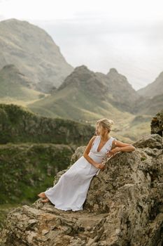 Woman in white dress relaxing on a mountain cliff with stunning Greek landscape backdrop.