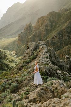 Woman in white dress walking along rocky mountain path in Greece during daylight.