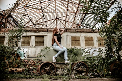 Side view of young female in casual outfit touching long hair while sitting on old rustic carriage in greenhouse