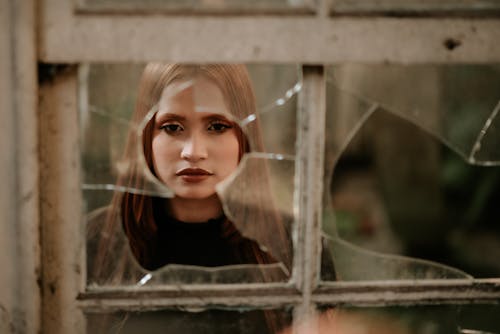 Спокойная молодая женщина смотрит в камеру через разбитое окно