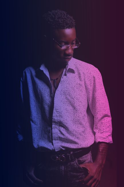 Dreamy young African American guy in stylish outfit and eyeglasses standing with hands on pockets and looking down under purple neon light in dark room