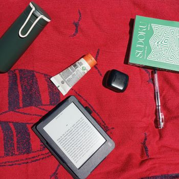 Flat lay of a summer beach setup with an e-reader, Sudoku book, and essentials on a towel.