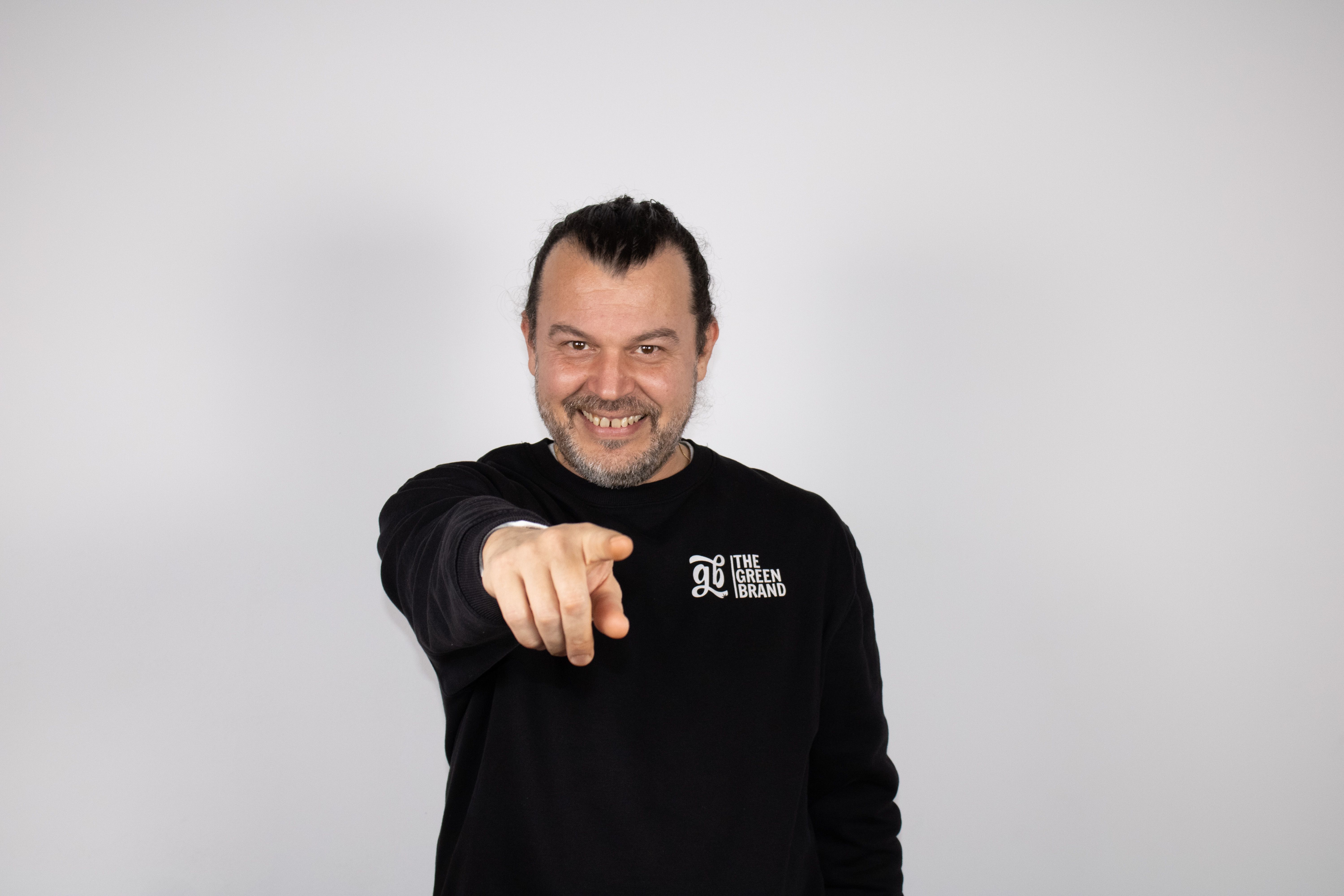 Cheerful man in black shirt pointing at camera against a white background, conveying engagement.