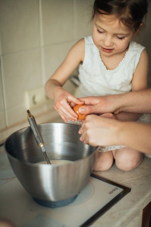 Free A young girl cracks an egg into a mixing bowl, learning to bake with guidance. Stock Photo