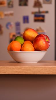 Fresh apples, oranges, and limes in a white bowl on a wooden counter in a home kitchen.