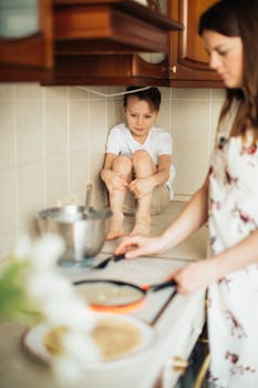 小男孩坐在柜台上，他母亲在厨房里舒适地做饭