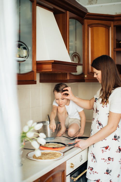 Cheerful woman in apron cooking pancakes while son sitting on kitchen cupboard near cooker and pointing to crepe