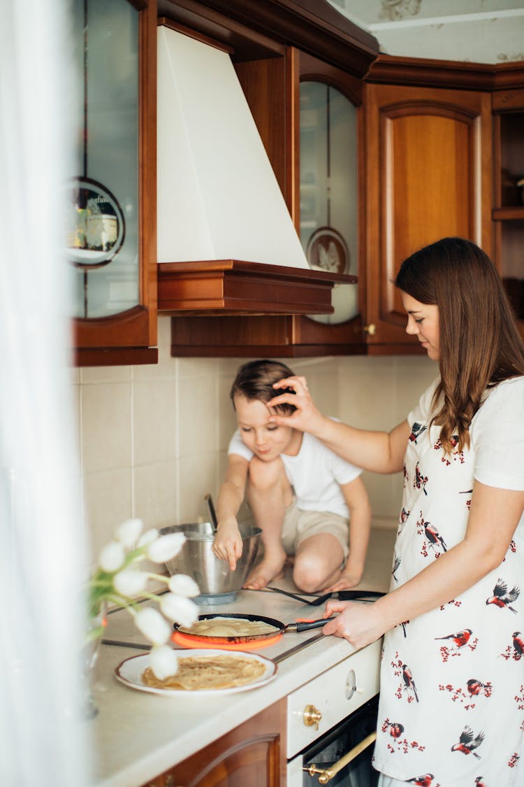 Young Mother With Son Cooking Crepes In Kitchen