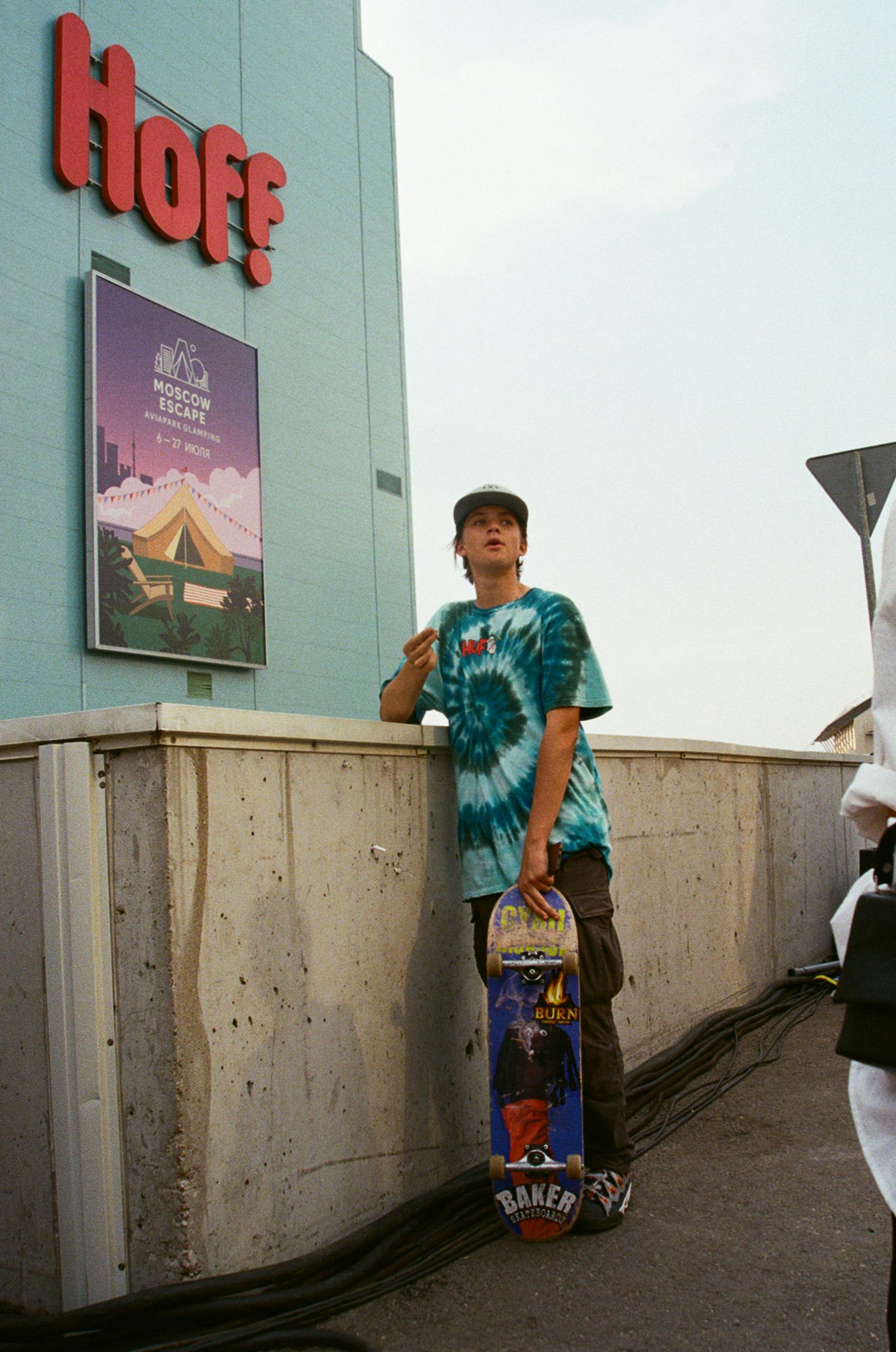 A young skateboarder in tie-dye shirt next to a Moscow-themed poster outdoors.