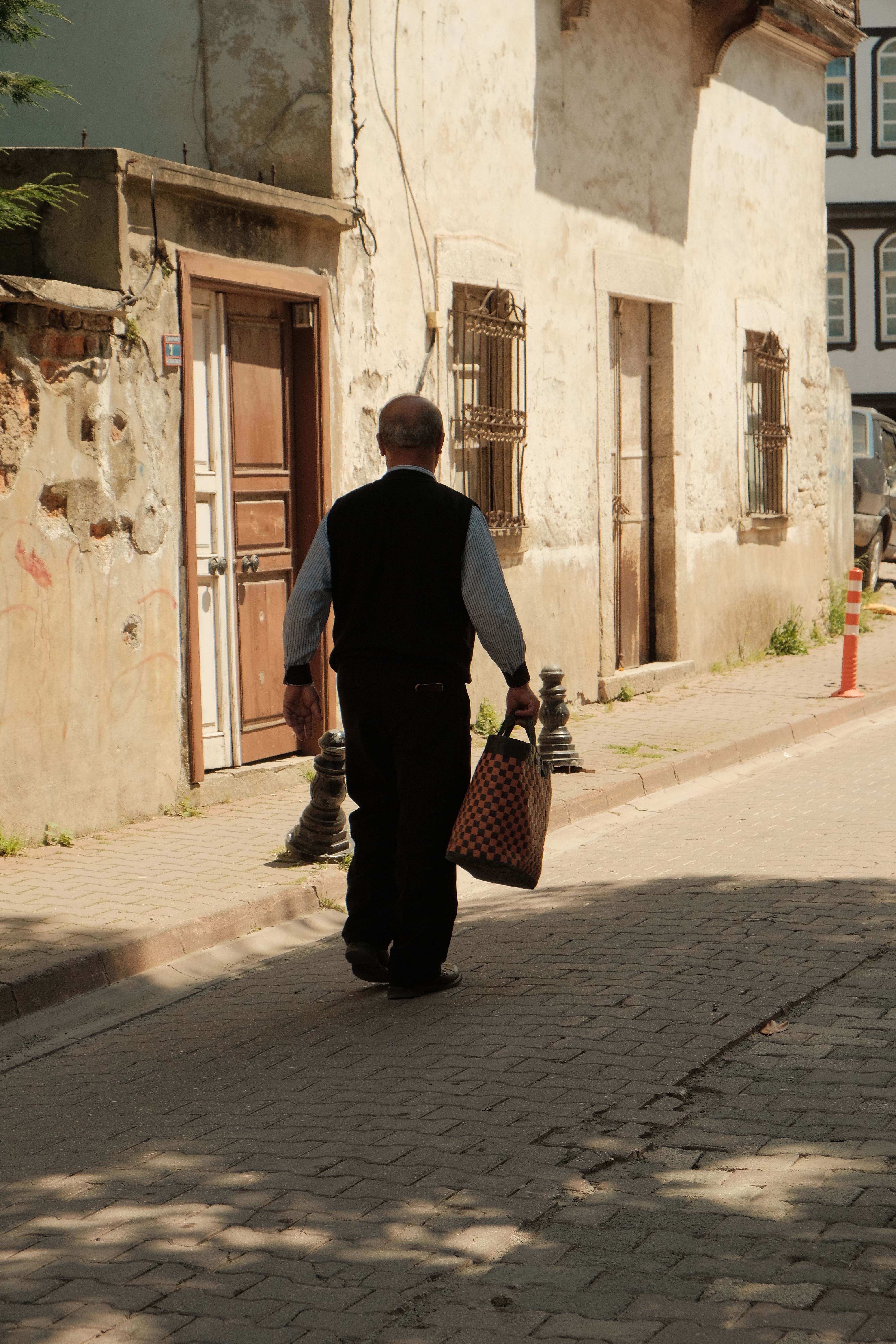 An elderly man carries a woven bag as he strolls down a sunlit historic street.