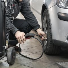 Mechanic crouching by a silver car in a bright workshop, checking the tyre with a round-headed gauge on a hose