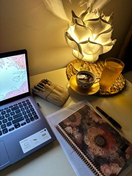Warmly lit desk setup featuring a laptop, notebooks, and a decorative lamp.